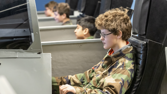 A young trainee wearing camouflage fatigues sits in the cockpit of an flight simulator. His team can be seen seated in their own sims beside him.