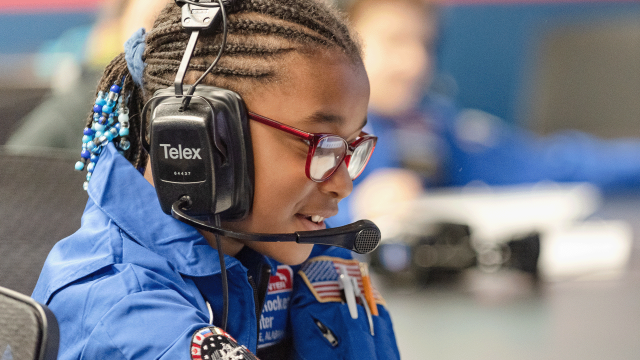 A young camper wears an adult sized headset and speaks into the attached microphone from Mission Control during a space flight simulated